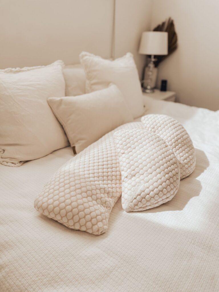 A soft, textured pregnancy pillow rests on a neatly made bed with cream-colored pillows and bedding. The serene setup emphasizes the role of supportive pillows in promoting restful sleep, essential for postpartum recovery.